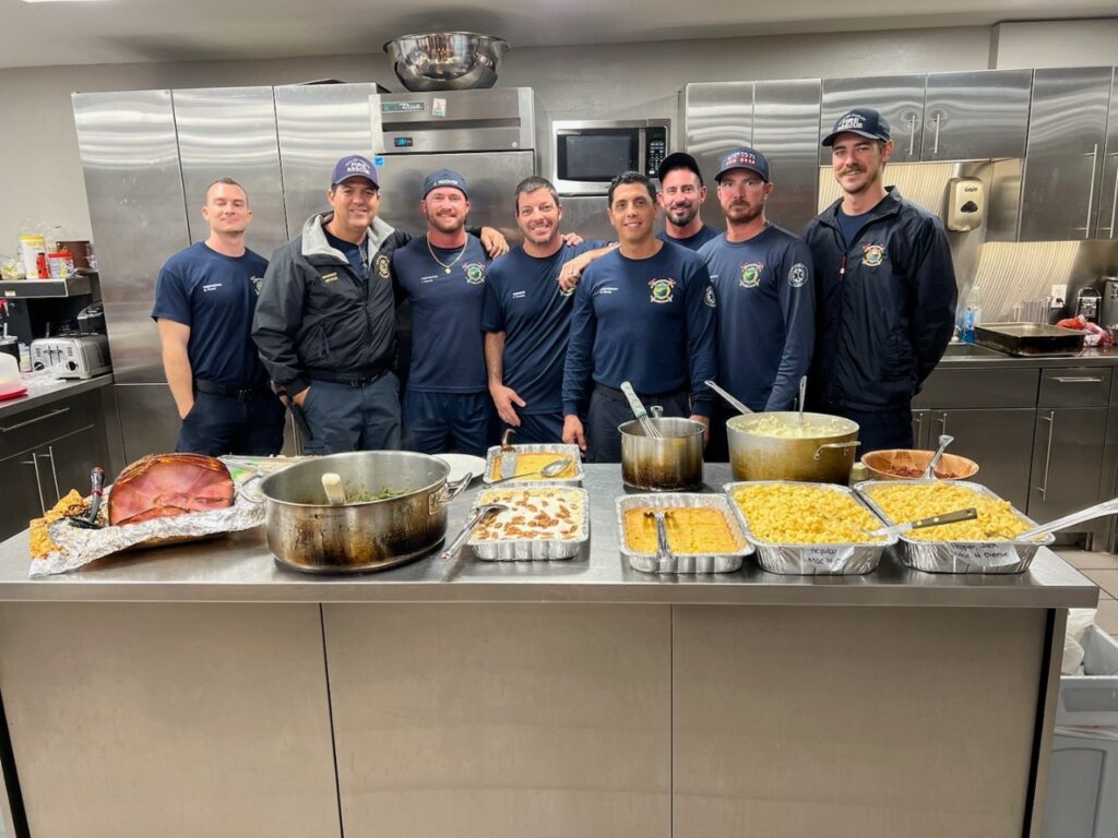 A group of firefighters in a fire station having a potluck. They are embracing each other and discussing going on a first responder retreat for mental health.