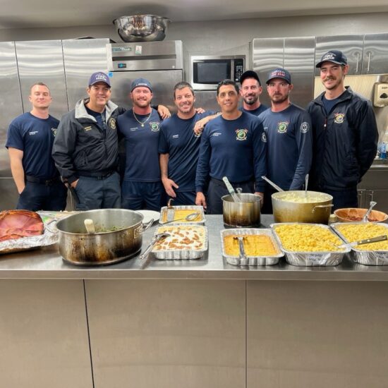 A group of firefighters in a fire station having a potluck. They are embracing each other and discussing going on a first responder retreat for mental health.