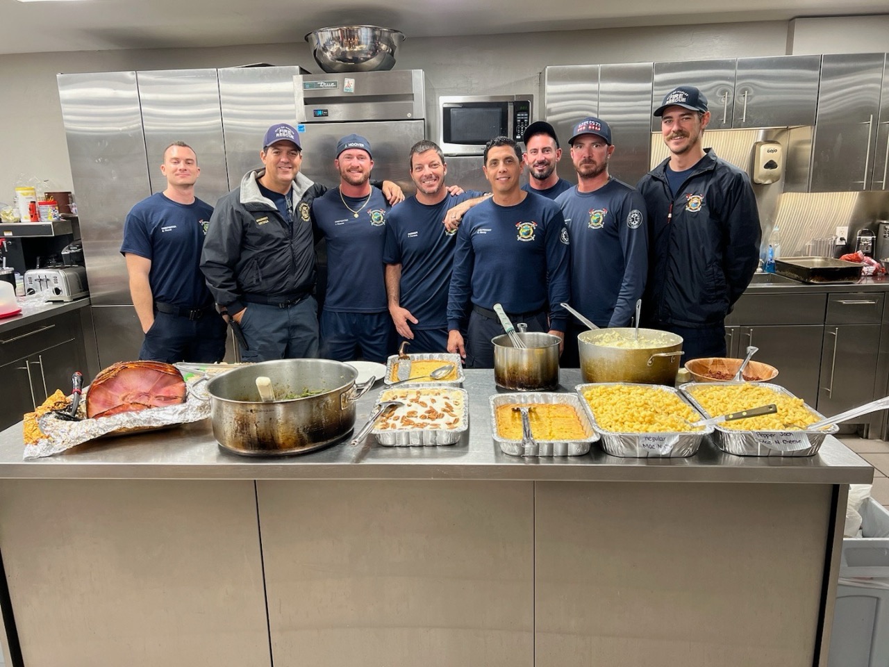 A group of firefighters in a fire station having a potluck. They are embracing each other and discussing going on a first responder retreat for mental health.