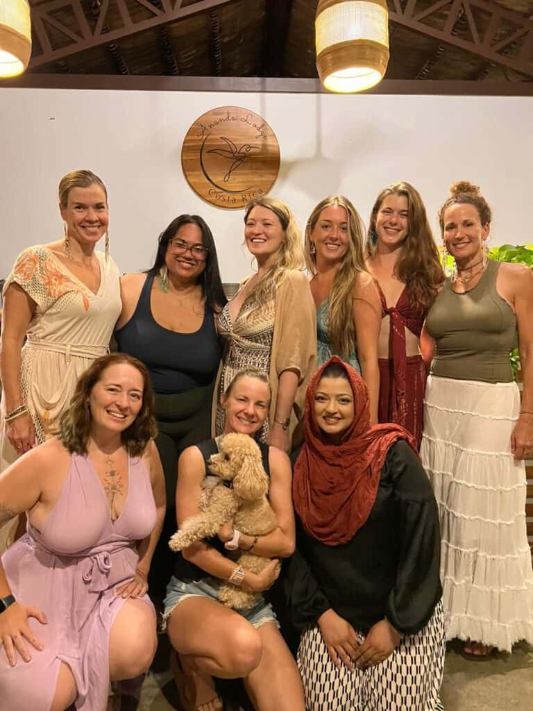 Group of nine women smiling and embracing at one of Ananda Lodge's women-only somatic healing retreats, with a friendly dog beside them, all illuminated by soft, natural light that conveys warmth and connection.