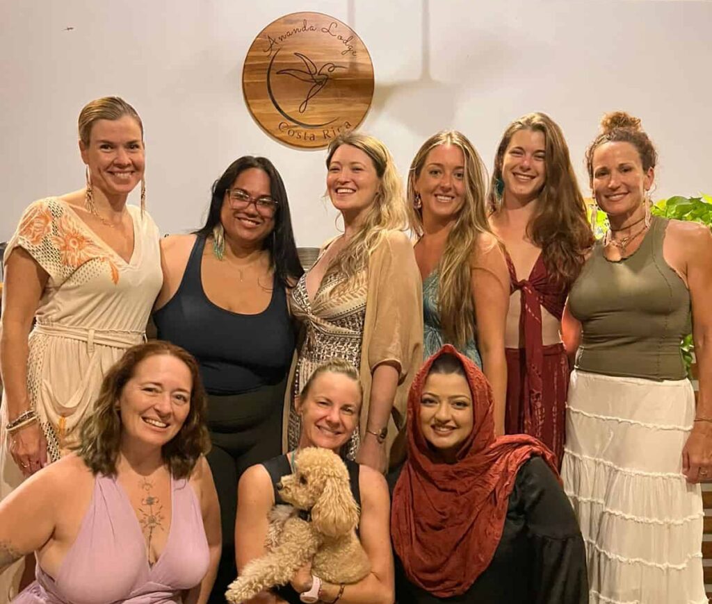 Group of nine women smiling and embracing at one of Ananda Lodge's women-only somatic healing retreats, with a friendly dog beside them, all illuminated by soft, natural light that conveys warmth and connection.