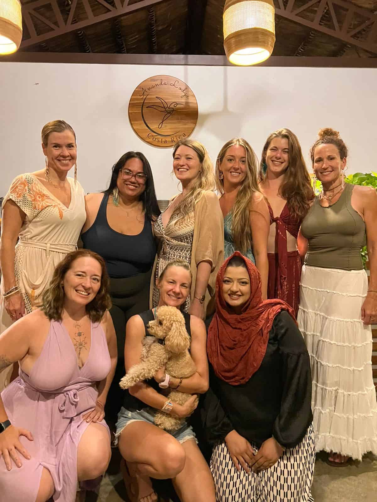 Group of nine women smiling and embracing at one of Ananda Lodge's women-only somatic healing retreats, with a friendly dog beside them, all illuminated by soft, natural light that conveys warmth and connection.
